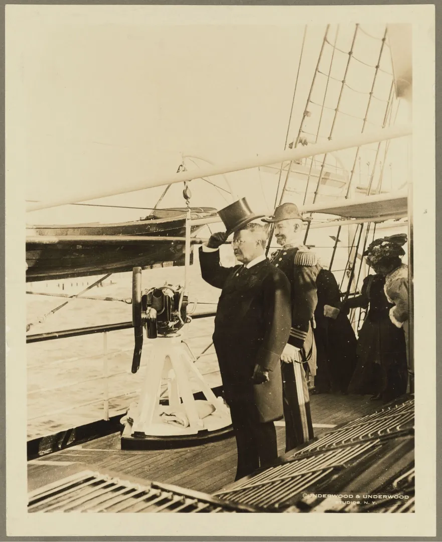 President Theodore Roosevelt in a top hat waving from the deck of a U.S. battleship during a naval review