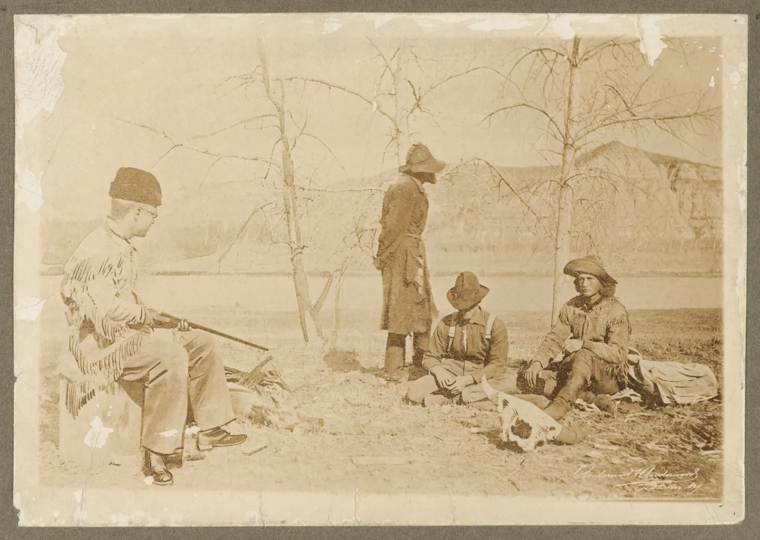 Men seated on a Badlands riverbank while one figure stands guard with a rifle during the 1886 pursuit of boat thieves.