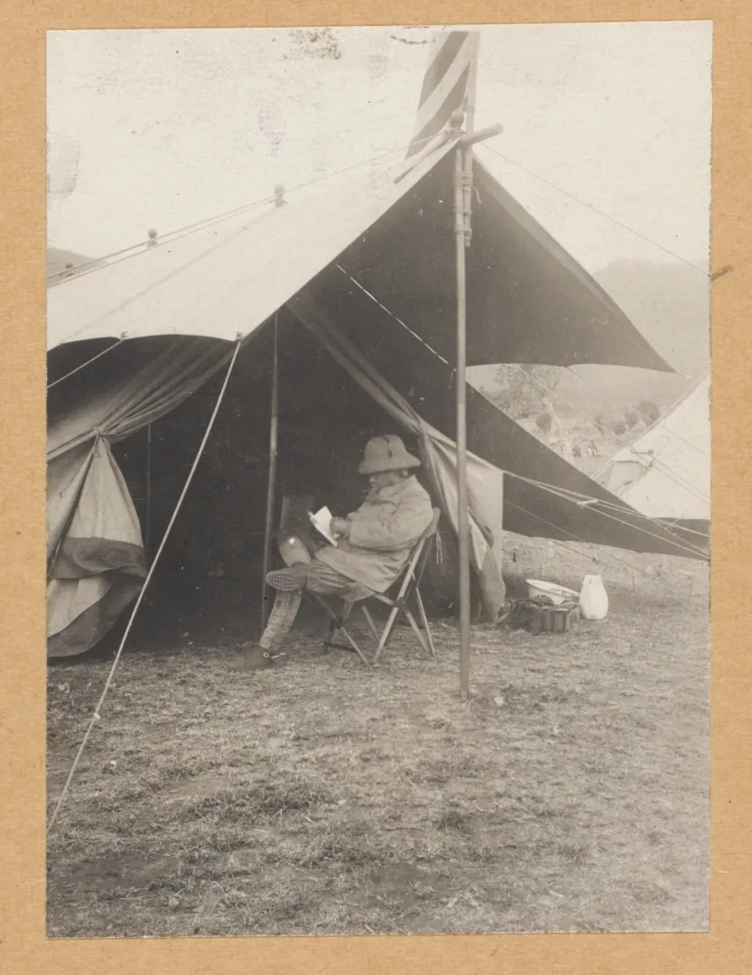 Theodore Roosevelt reading while seated in front of a tent at a wilderness camp