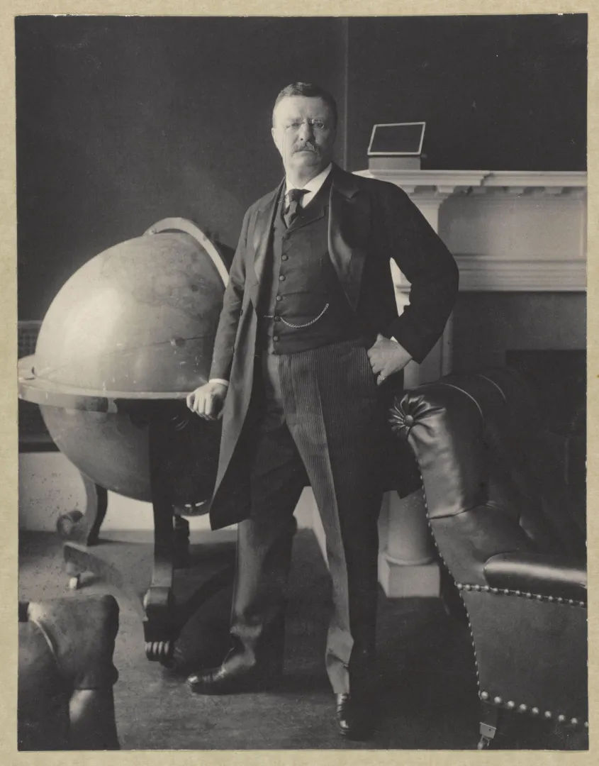 President Theodore Roosevelt in a full-length studio portrait, standing beside a large globe