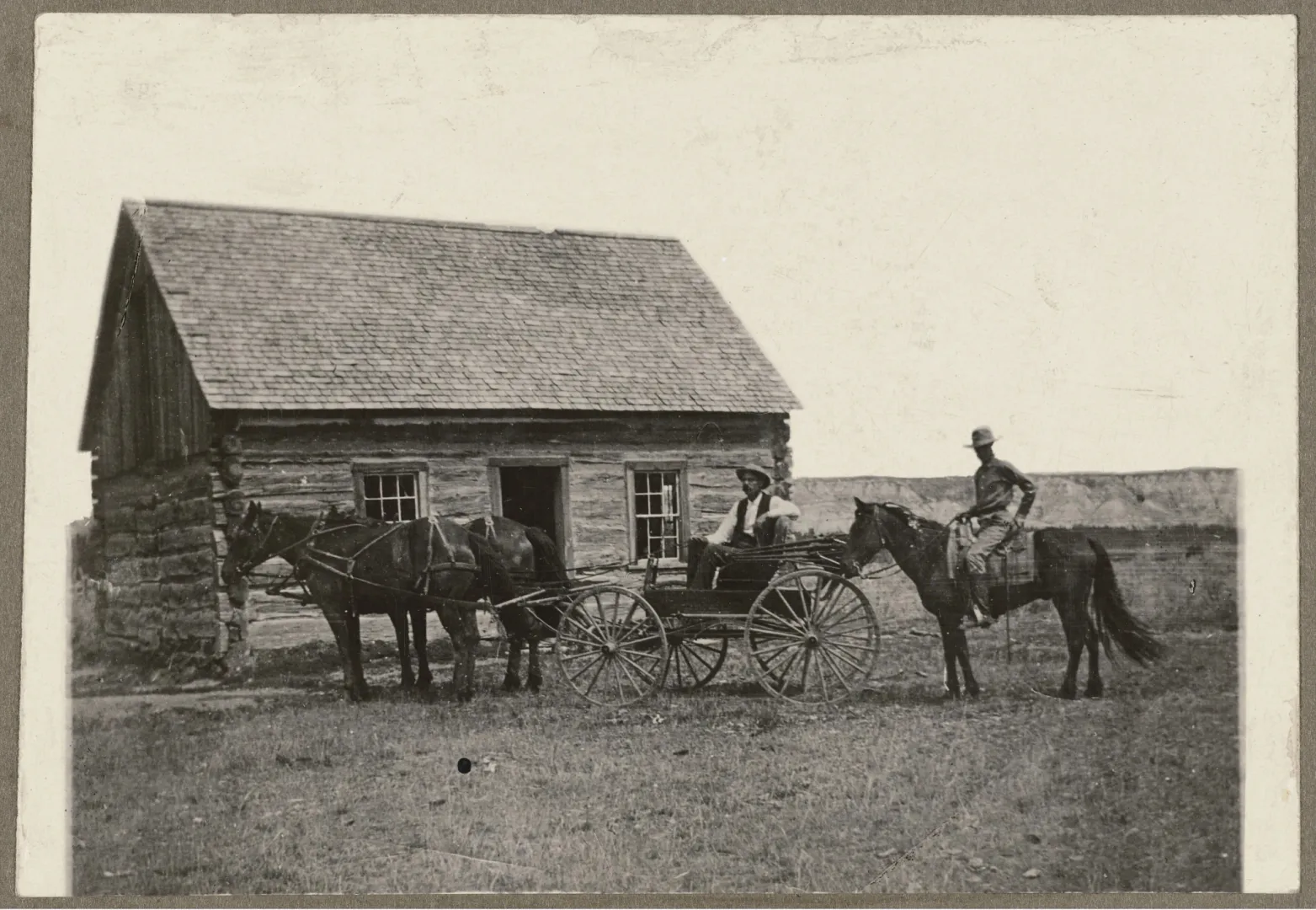 Historic photograph of the Maltese Cross Ranch cabin, a low log house with a shingled roof, with a team of horses and two mounted riders in front