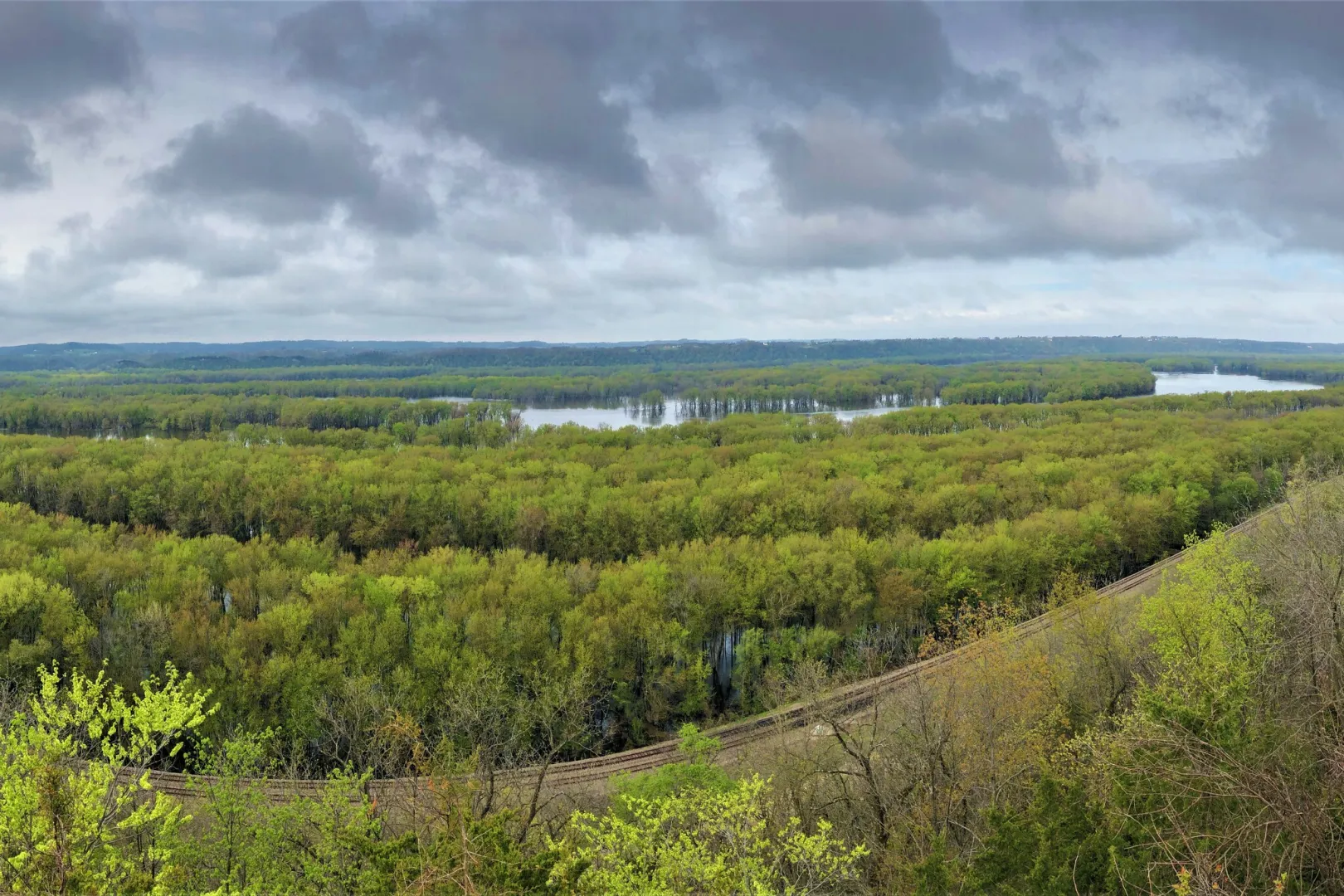 View from a bluff over the wooded floodplain and open water of the Upper Mississippi River under a cloudy sky