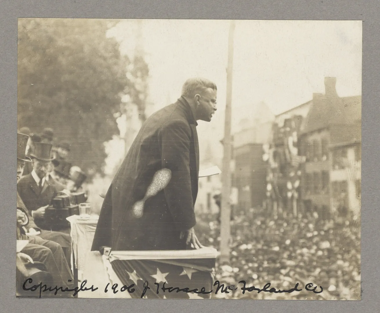 Theodore Roosevelt leaning forward to address a large crowd from a flag-draped platform