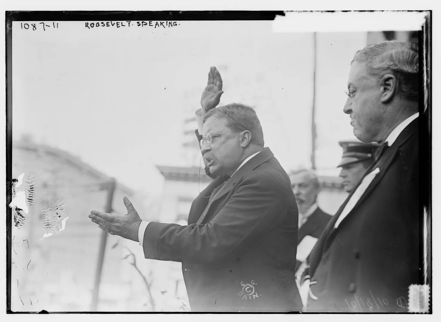 Theodore Roosevelt raising his hand while delivering a speech beside another man