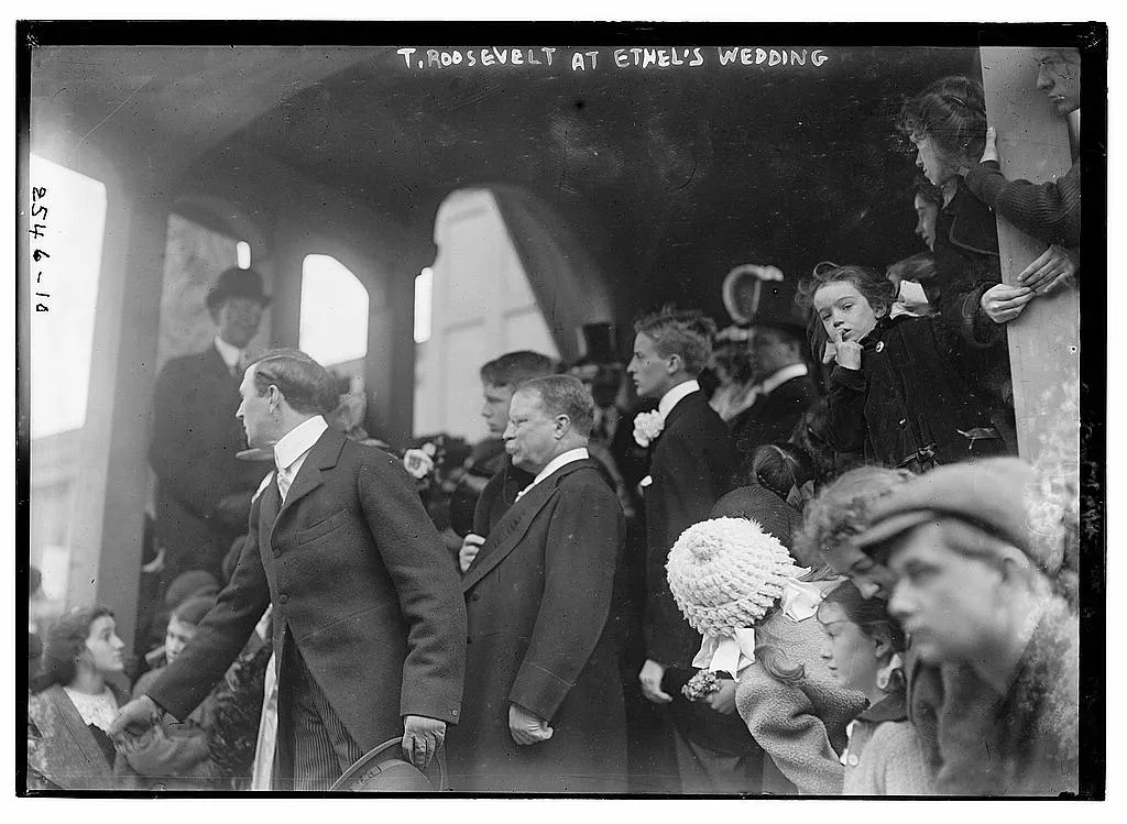 Theodore Roosevelt in a dark overcoat standing among wedding guests under a covered porch, 1913
