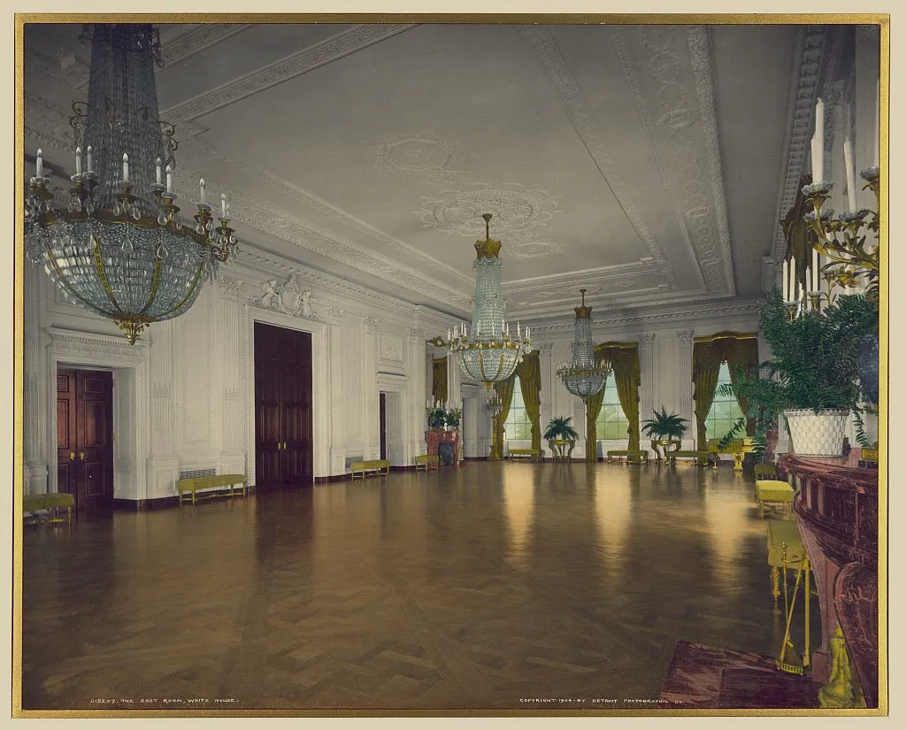 A long white ballroom with three crystal chandeliers, tall windows with gold curtains and a gleaming parquet floor.