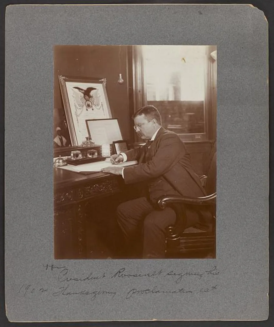 President Theodore Roosevelt seated at a desk with a pen, signing a document