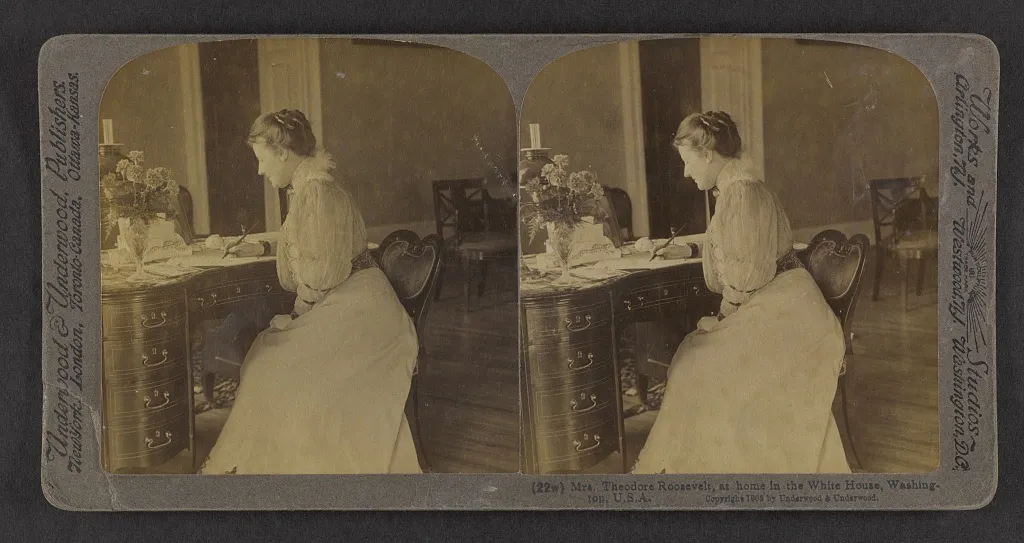 Stereograph card showing Edith Roosevelt in a pale blouse and long skirt, writing at a desk with a vase of flowers and a candle.