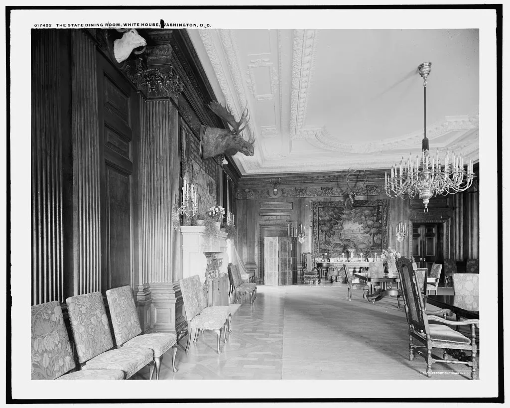 A long oak-paneled dining room with mounted moose and sheep heads high on the walls, a tapestry at the far end, a branched chandelier and tapestry-covered chairs.