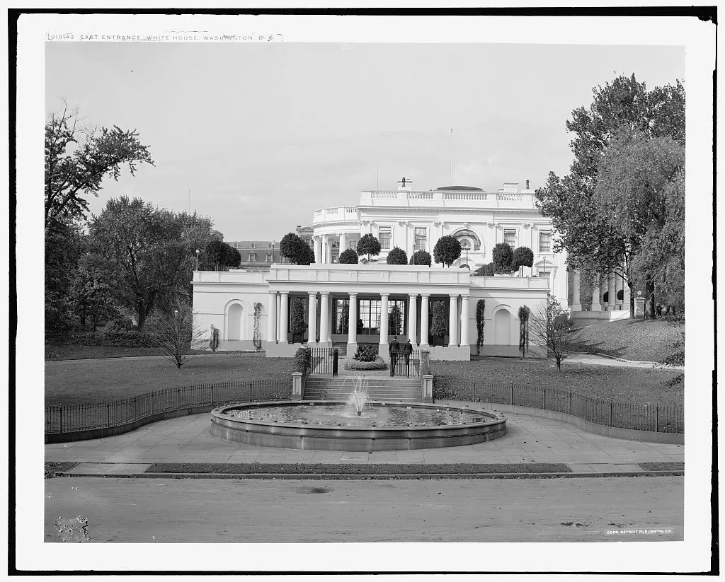 The low colonnaded east wing of the White House with a covered carriage entrance and a round fountain in front.