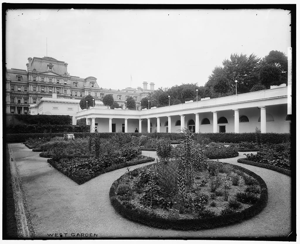 A formal garden of round and curving flower beds edged with low hedges, with the West Terrace colonnade and the State, War and Navy Building behind.