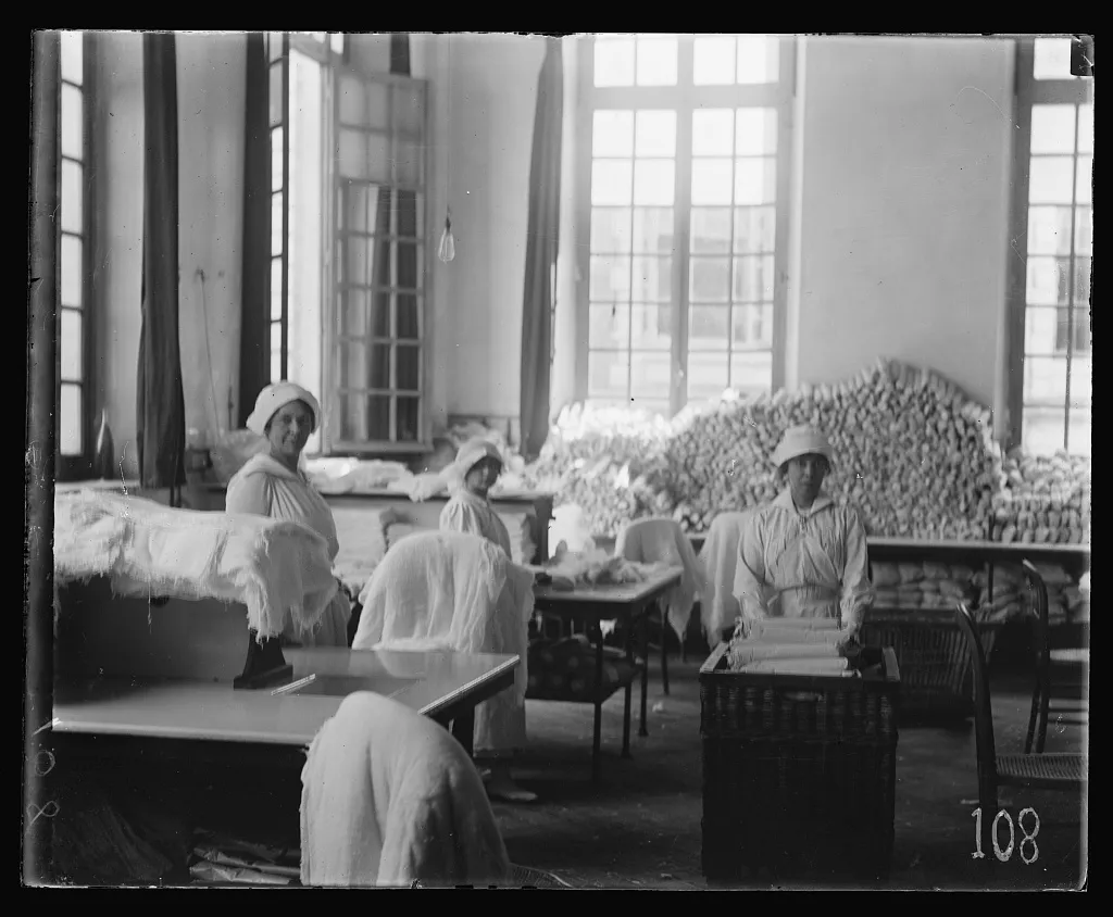 Three women in white caps and aprons at worktables in a tall-windowed room piled with rolled bandages and dressings.
