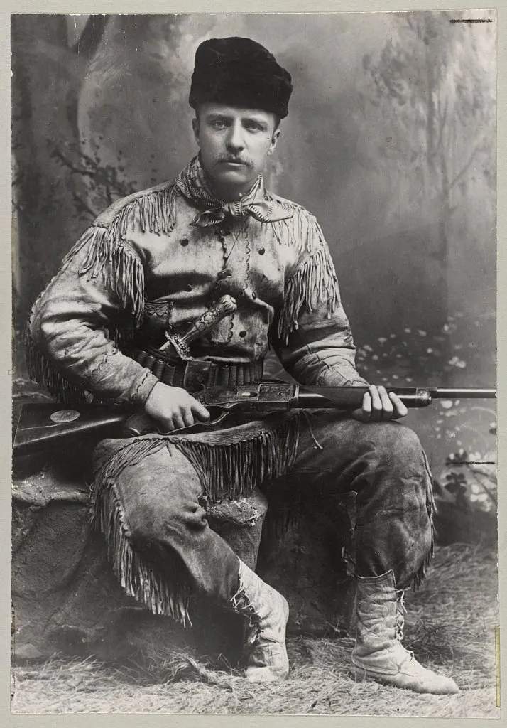 Studio portrait of young Theodore Roosevelt seated in a fringed buckskin hunting suit and fur cap, a rifle across his lap.