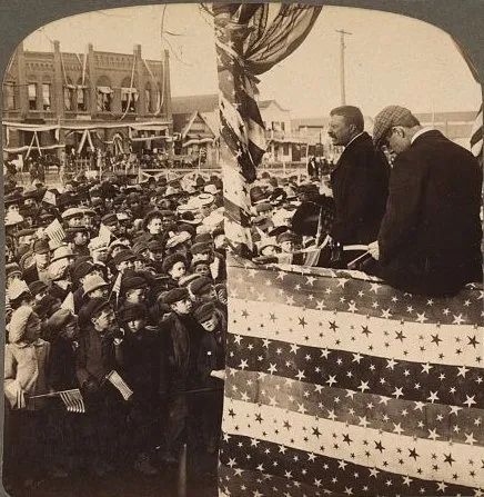 Theodore Roosevelt speaking from a flag-draped stand above a dense crowd, brick storefronts behind.
