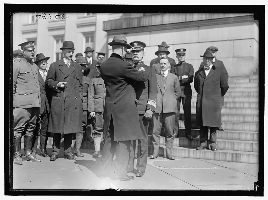 A naval officer with a white mustache, in dress uniform, has a medal pinned on his coat as a group of men looks on
