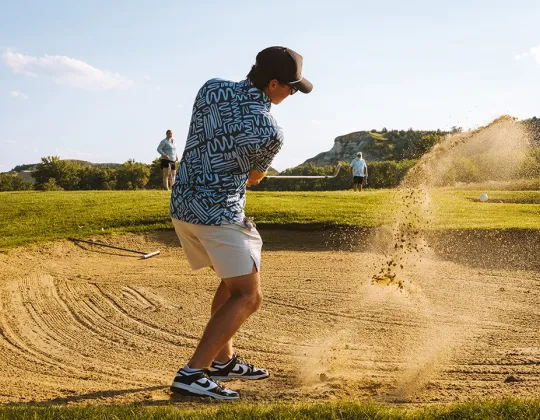 Golfer hitting out of a bunker with the Badlands behind